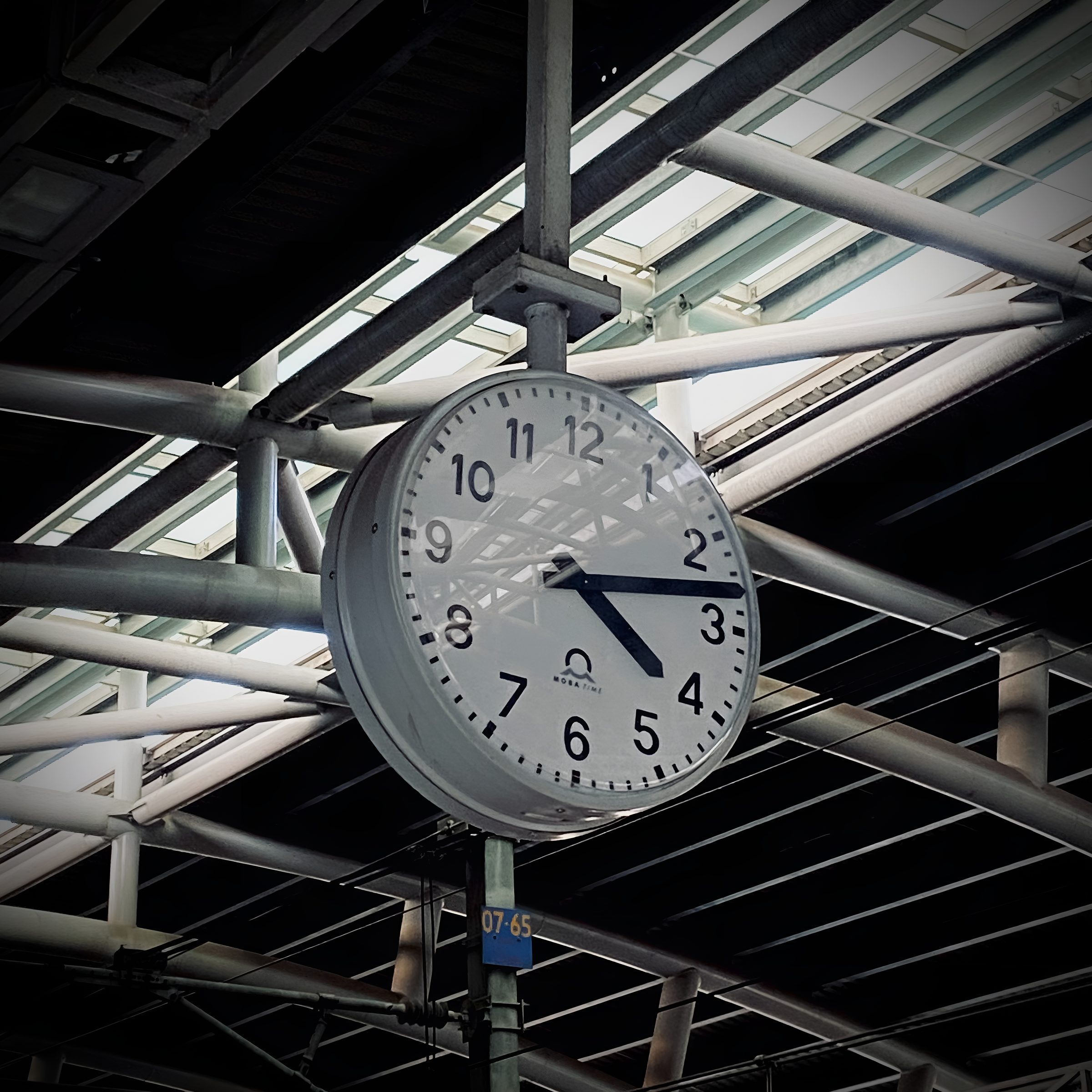 A large white station clock hanging from a steel beam under a glass roof